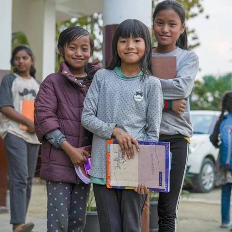 A group of four young girls stand outside holding schoolbooks and folders. Three of the girls are in the foreground, smiling at the camera.