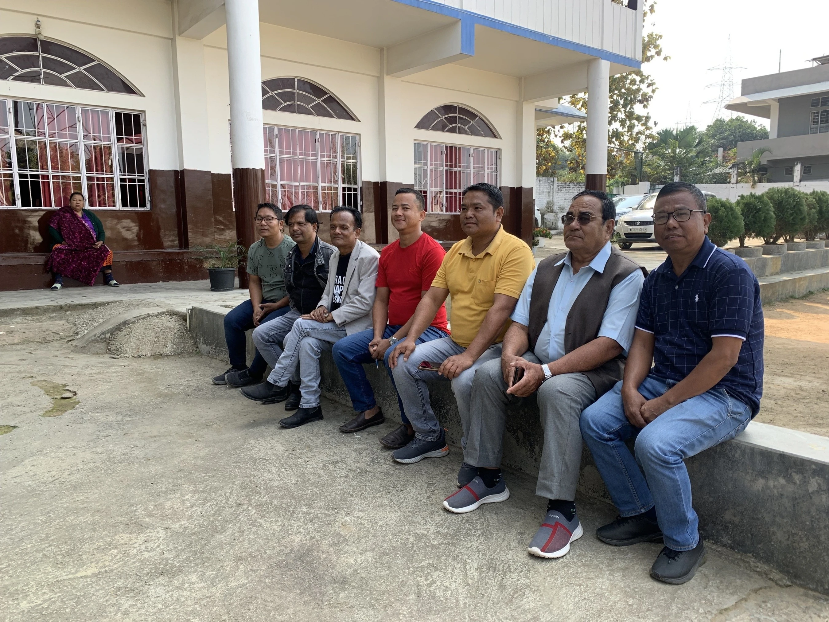 A group of eight men in casual clothing sit side-by-side on a concrete ledge in front of a building with large arched windows.