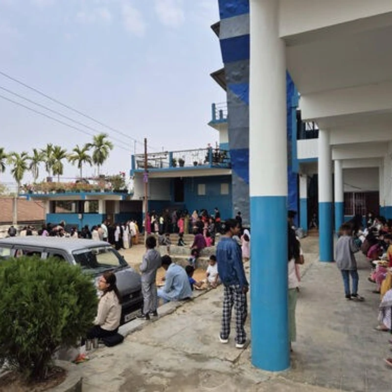 A large crowd of people gathered in the courtyard of a blue and white building. Some are eating along a covered walkway, while others stand and sit near a van.