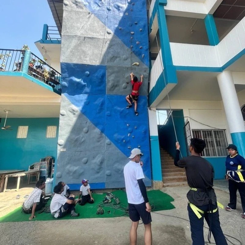A person in a red shirt climbs a tall blue and grey outdoor wall, while a group of people watch and belay from the ground on a sunny day.