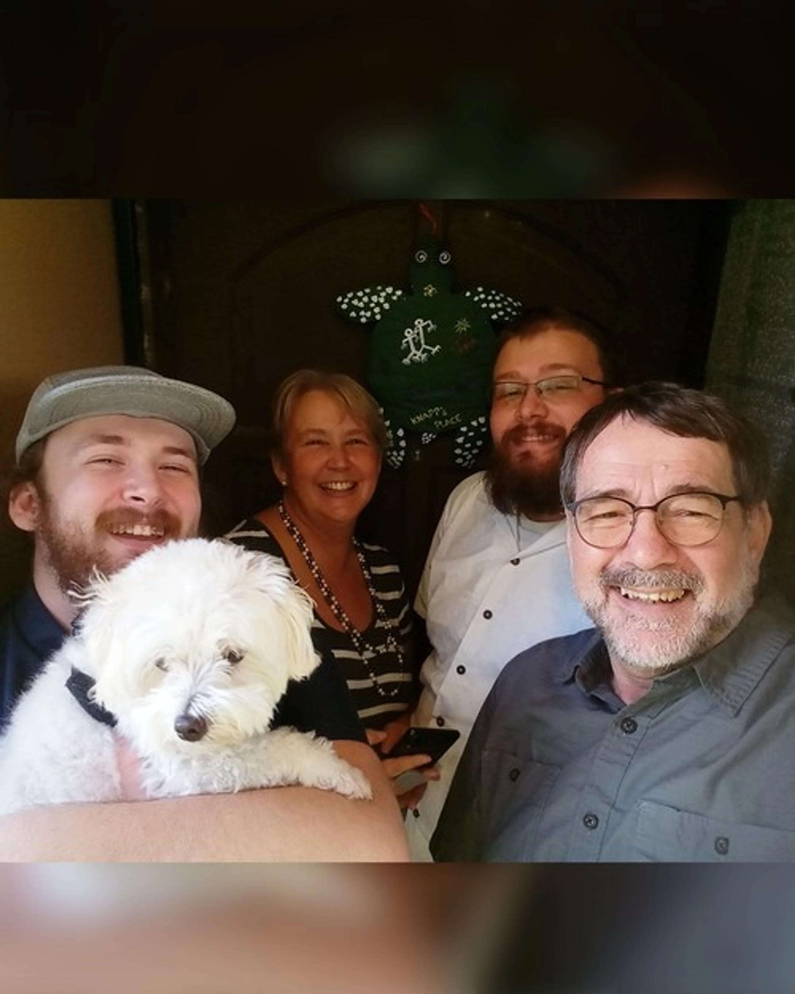 A group of four smiling people, three men and one woman, pose for a photo with a small, fluffy white dog. They are gathered closely together indoors.