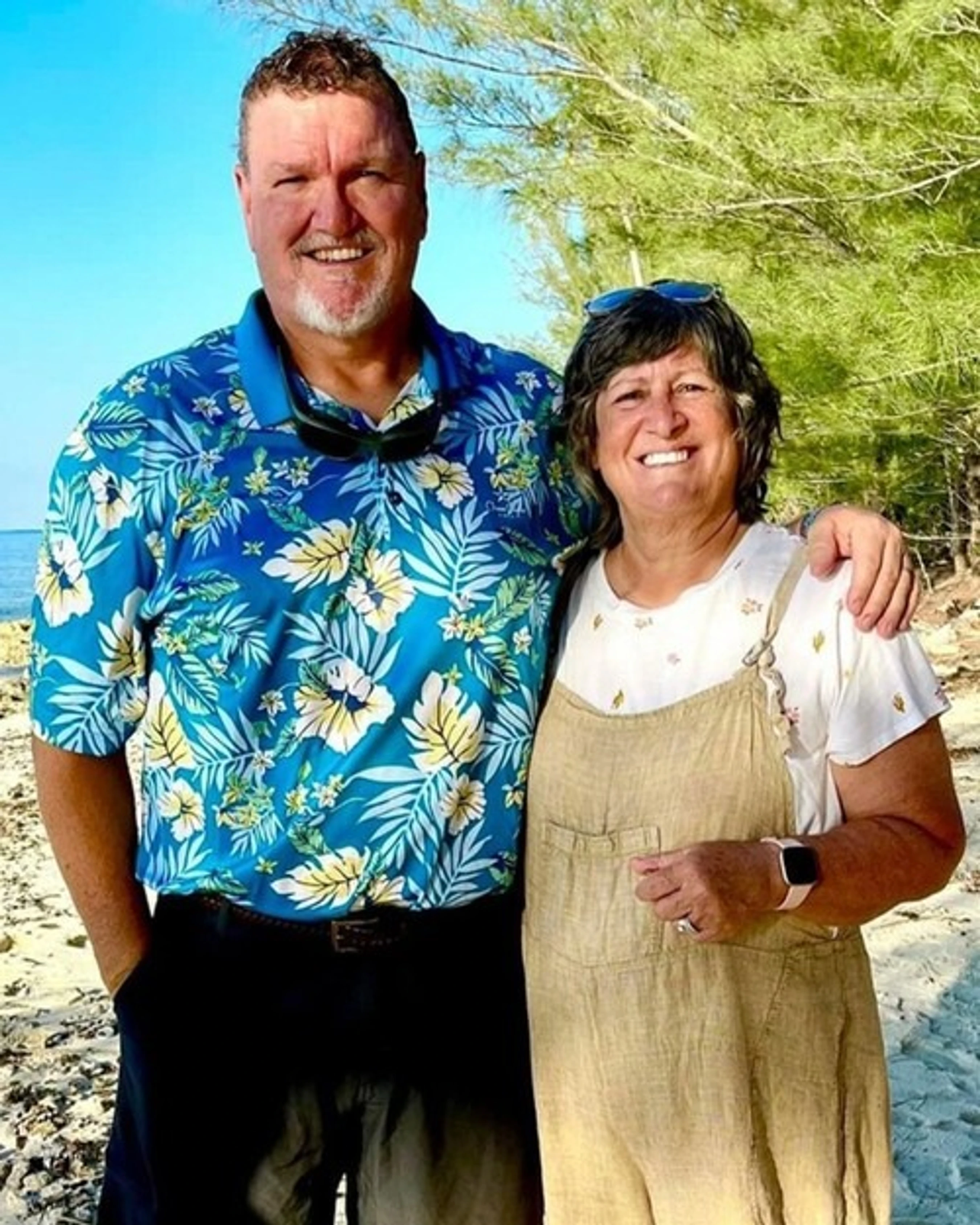 A smiling man in a blue floral shirt with his arm around a smiling woman in beige overalls. They are standing on a sunny beach with trees in the background.