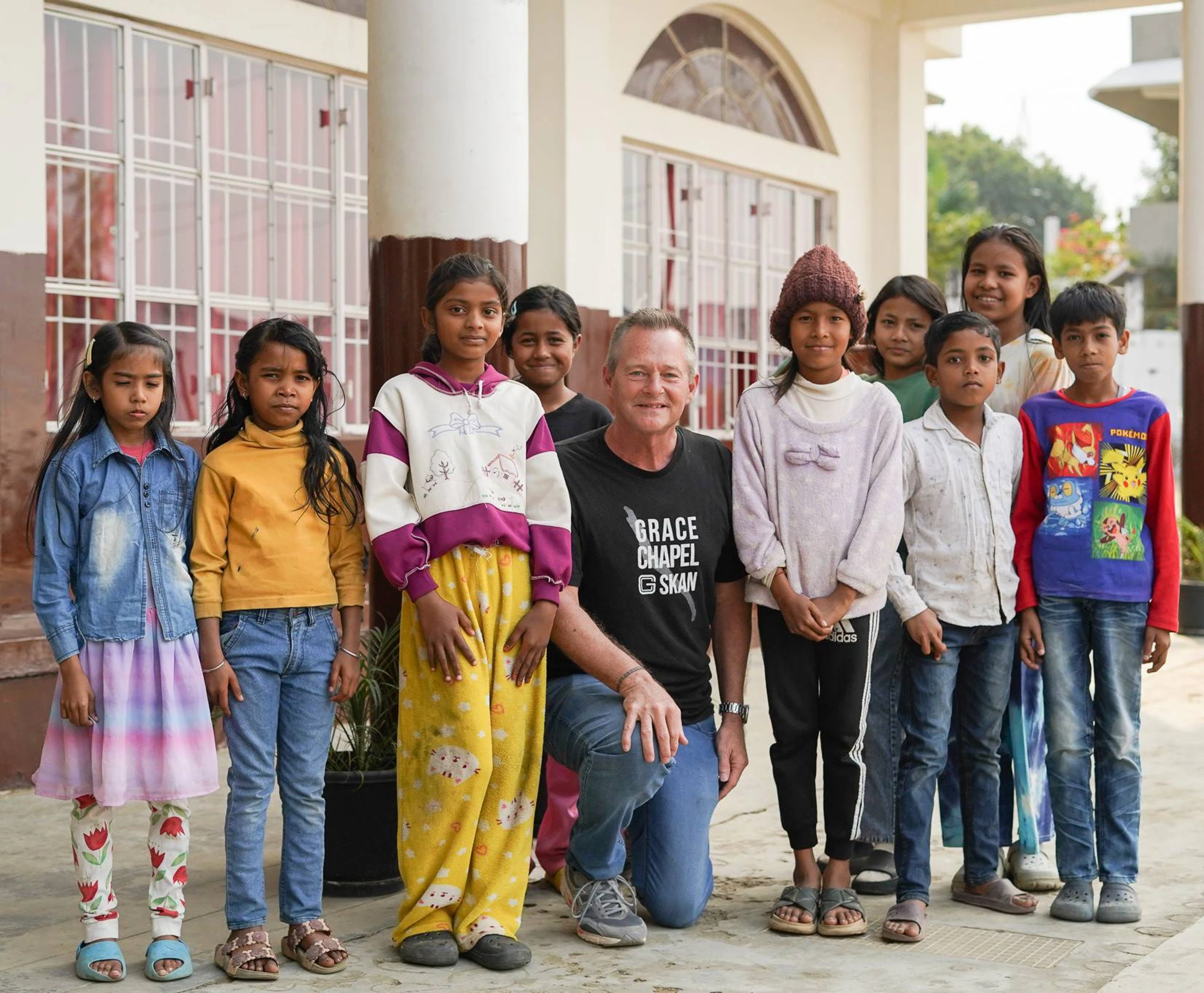 A man in a black Grace Chapel t-shirt kneels in the middle of a group of about ten children, posing for a photo in front of a school-like building.
