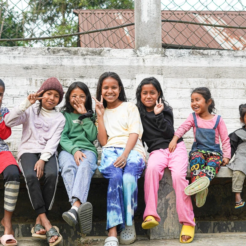 A group of seven young girls sitting in a row on a concrete ledge, smiling and making peace signs at the camera in front of a white wall and fence.