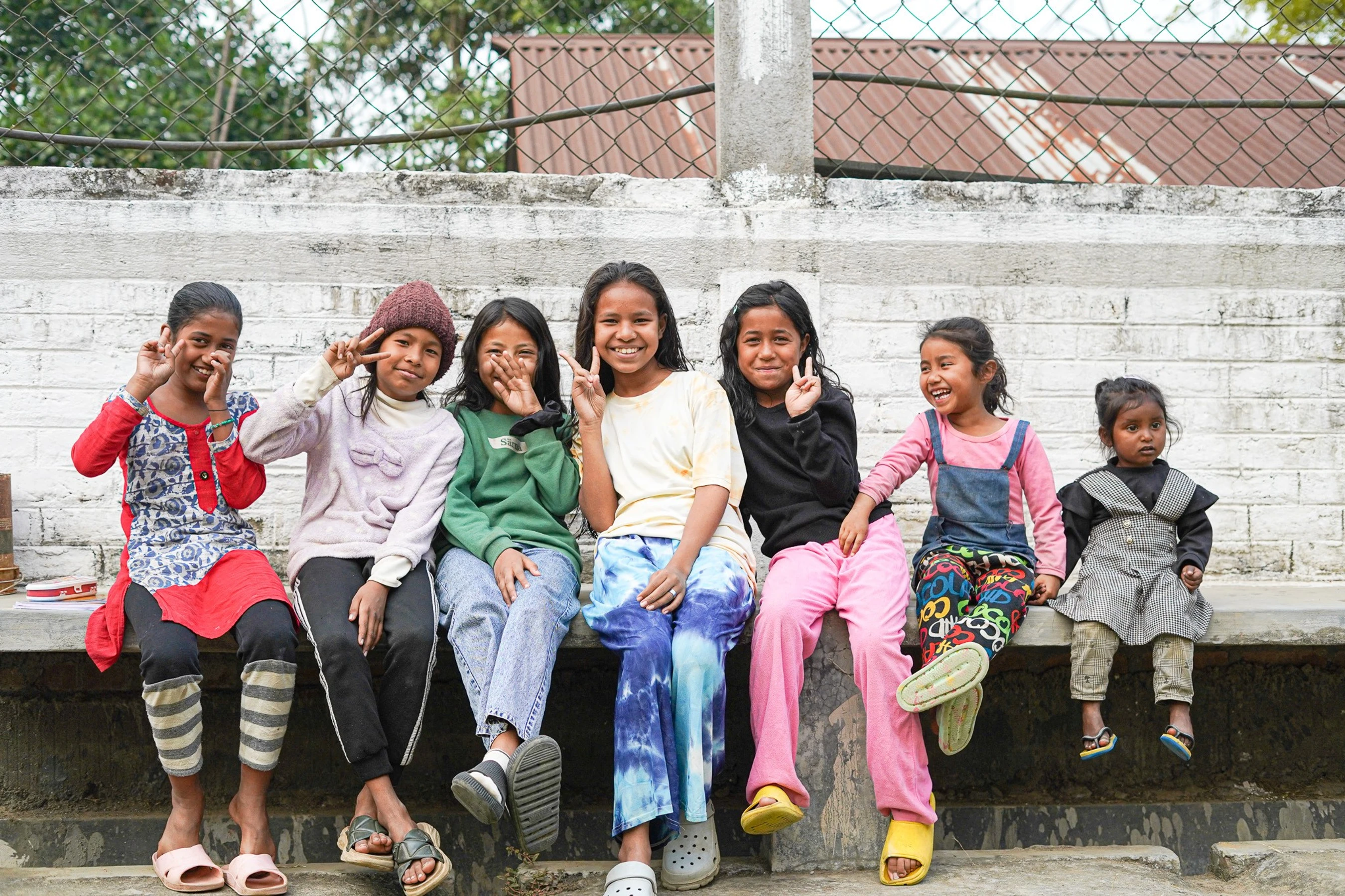 A group of seven young girls sitting in a row on a concrete ledge, smiling and making peace signs at the camera in front of a white wall and fence.