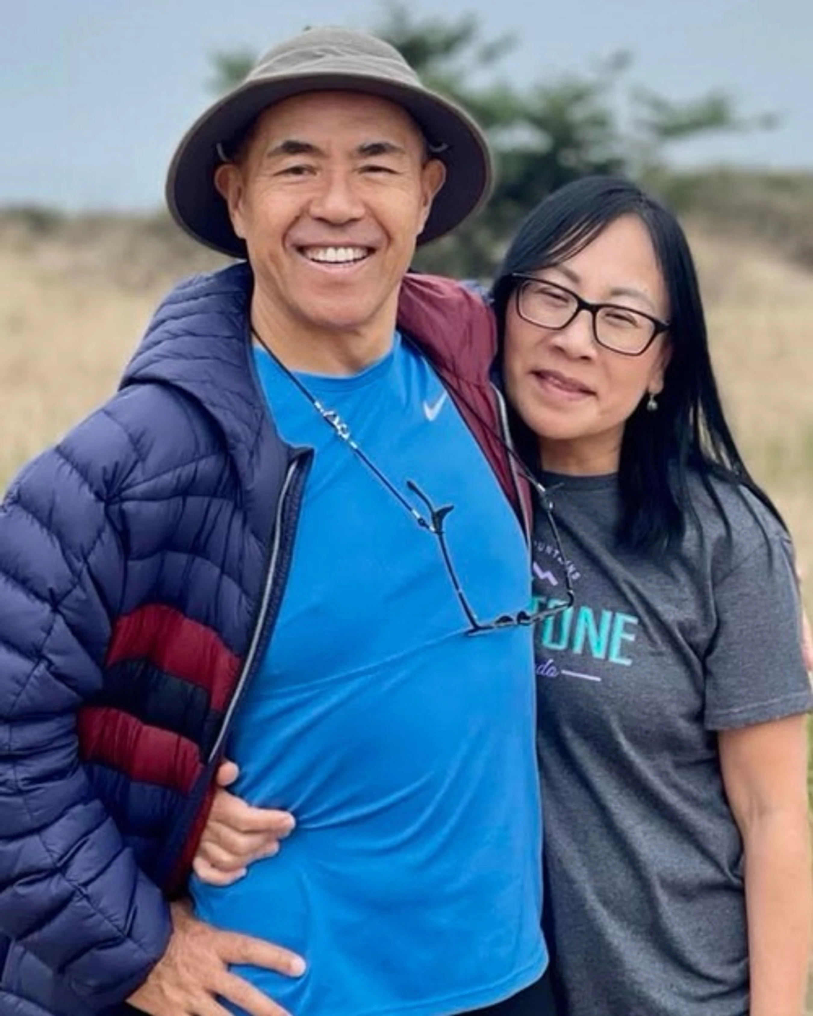 A smiling Asian man in a hat and blue jacket stands with his arm around a smiling Asian woman wearing glasses and a grey t-shirt in a natural setting.