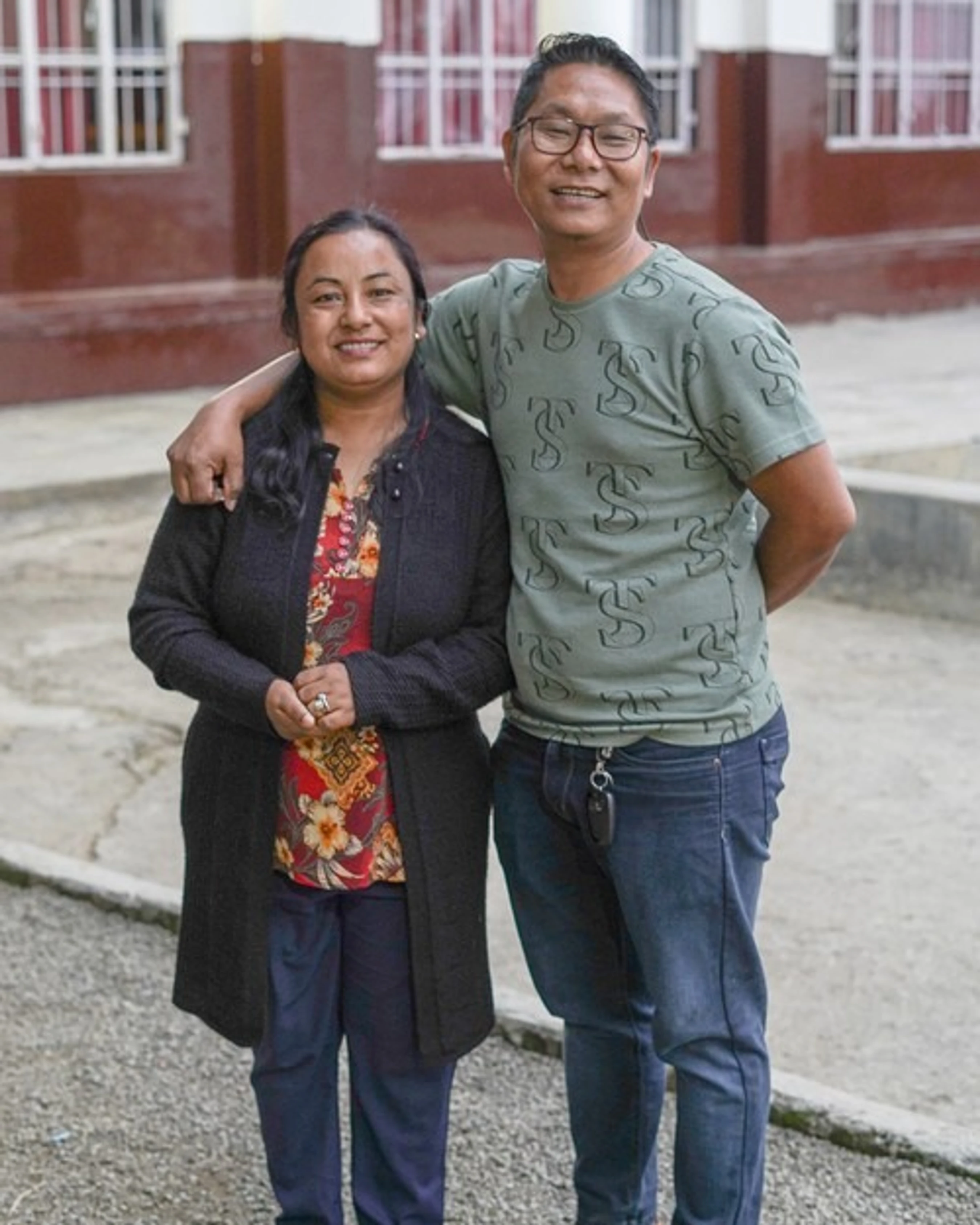 A man and a woman of Asian descent pose for a photo. The man has his arm around the woman, and both are smiling at the camera in an outdoor courtyard.