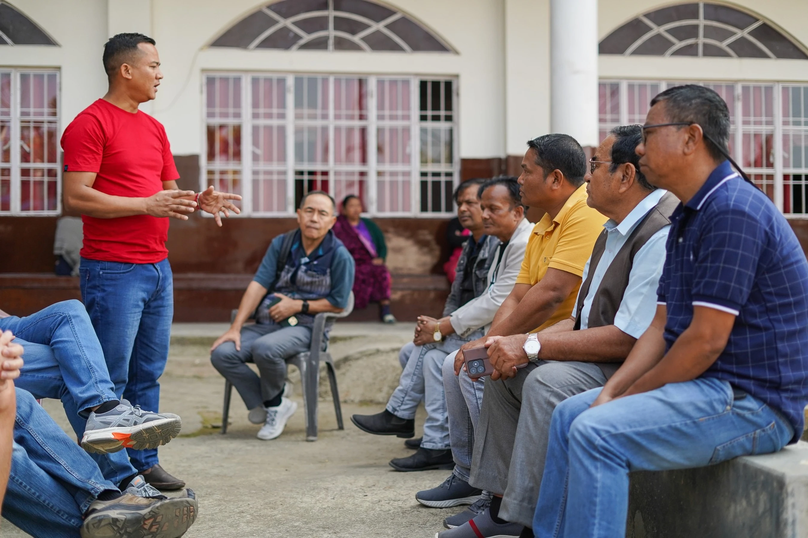 A man in a red t-shirt and jeans stands while speaking to a group of men sitting on a concrete ledge and listening attentively in front of a building.