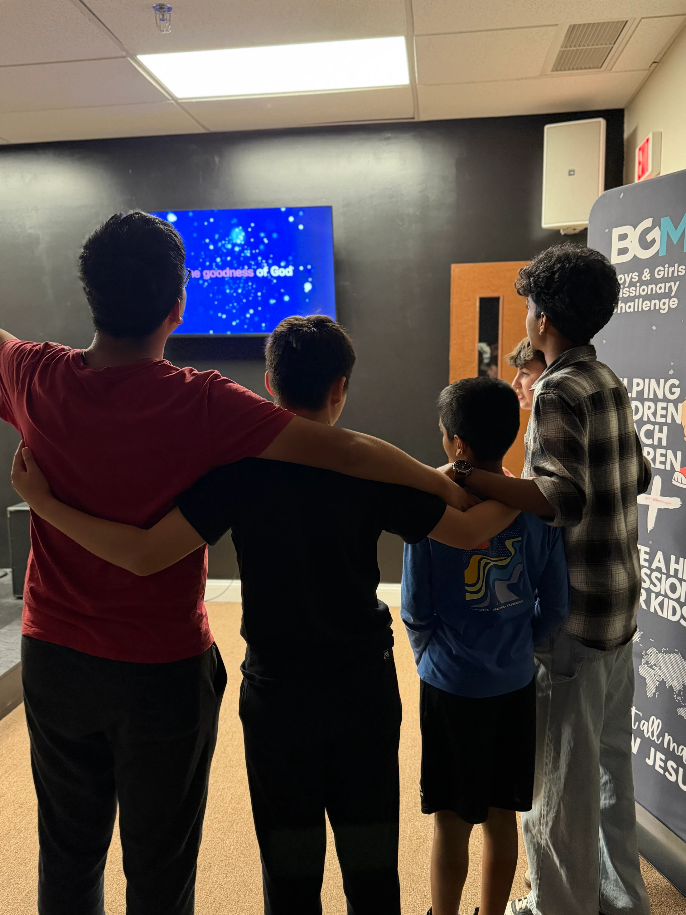 A group of four boys seen from behind with their arms around each other, looking at a screen with lyrics during a worship service.