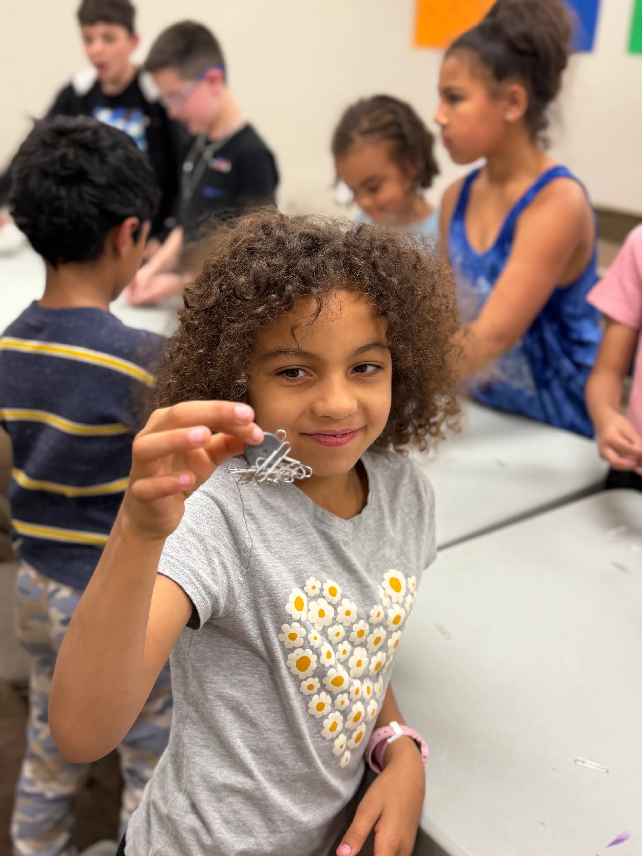 A smiling young girl with curly hair holds up a small creation made from paperclips. She is in a classroom with other children visible in the background.
