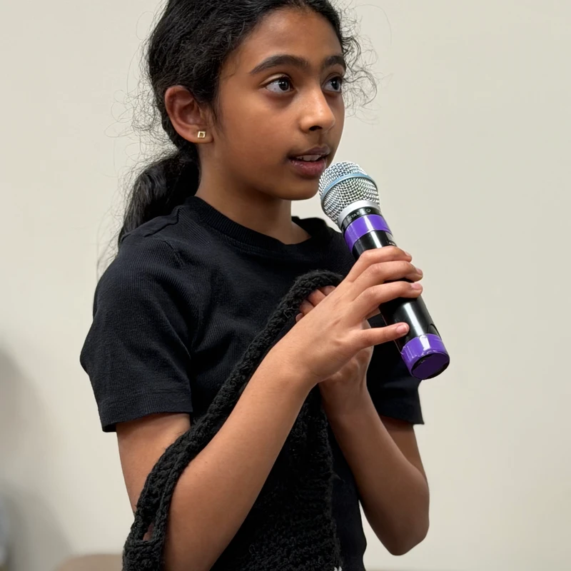 A young girl with dark curly hair tied back speaks into a microphone she holds with both hands. She wears a black shirt and a crocheted bag on her shoulder.