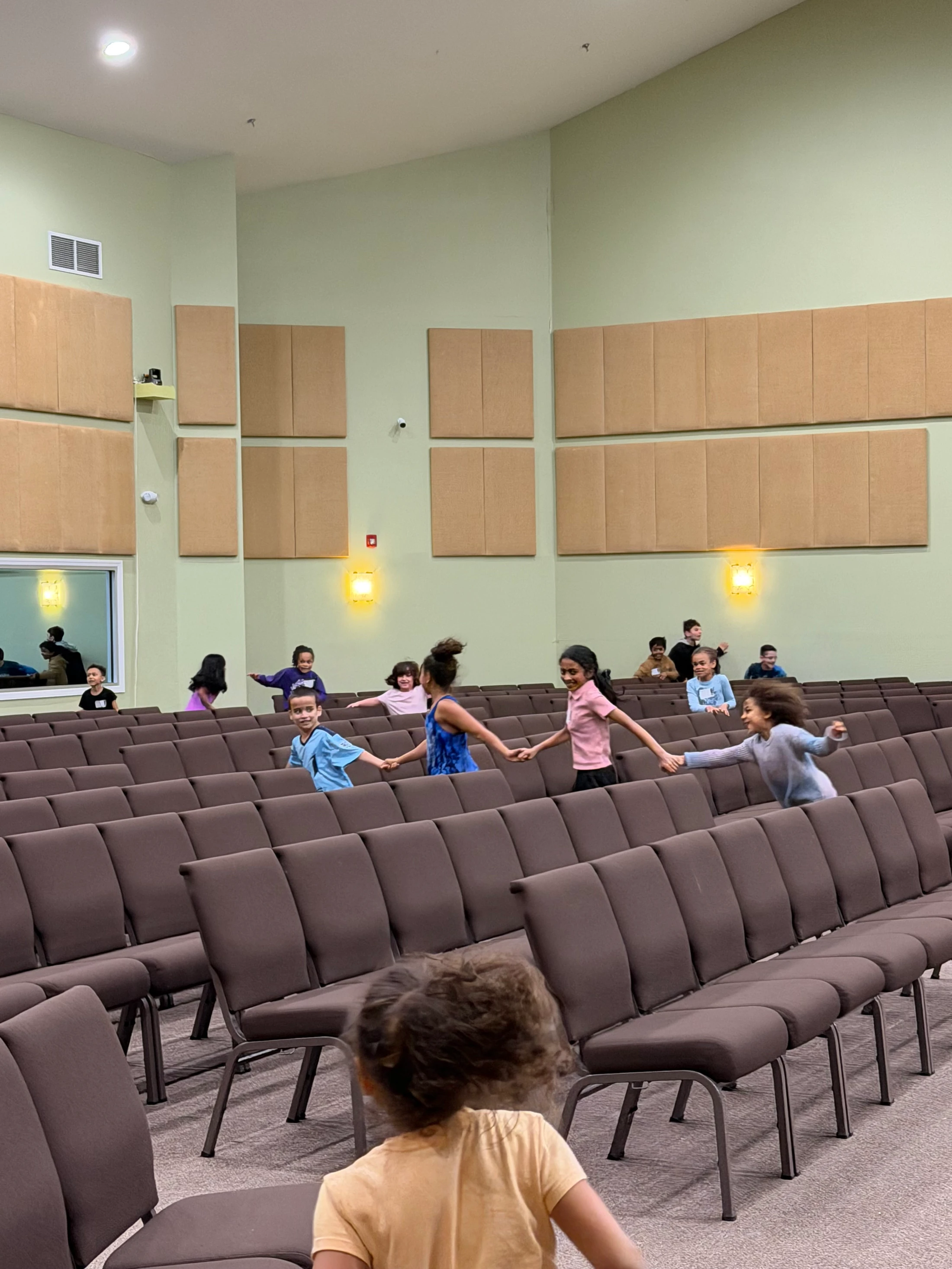 A group of children happily hold hands and run through the aisles of a large room filled with rows of brown padded chairs.
