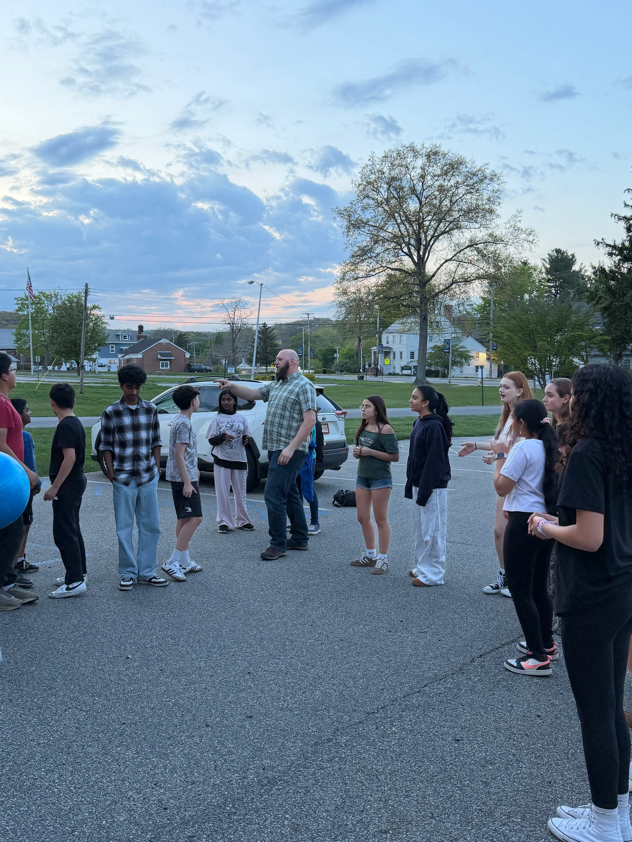 An adult man with a beard addresses a diverse group of teenagers standing in a circle in a parking lot during the evening, with a cloudy sky above.