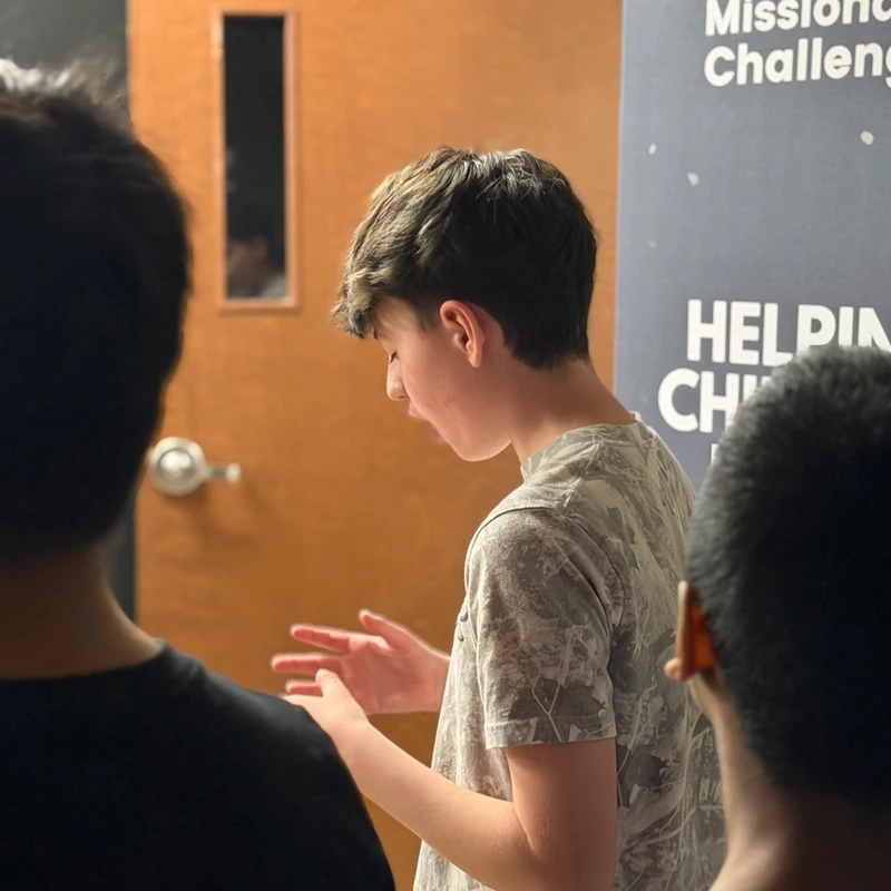 A young man in a camouflage t-shirt speaks and gestures with his hands while facing a small group, with a 'Boys & Girls Missionary Challenge' banner behind him.