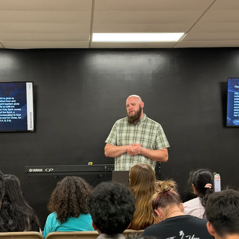 A bald man with a beard and a plaid shirt stands in front of a group of young people, speaking to them in a classroom-style setting with a black wall.