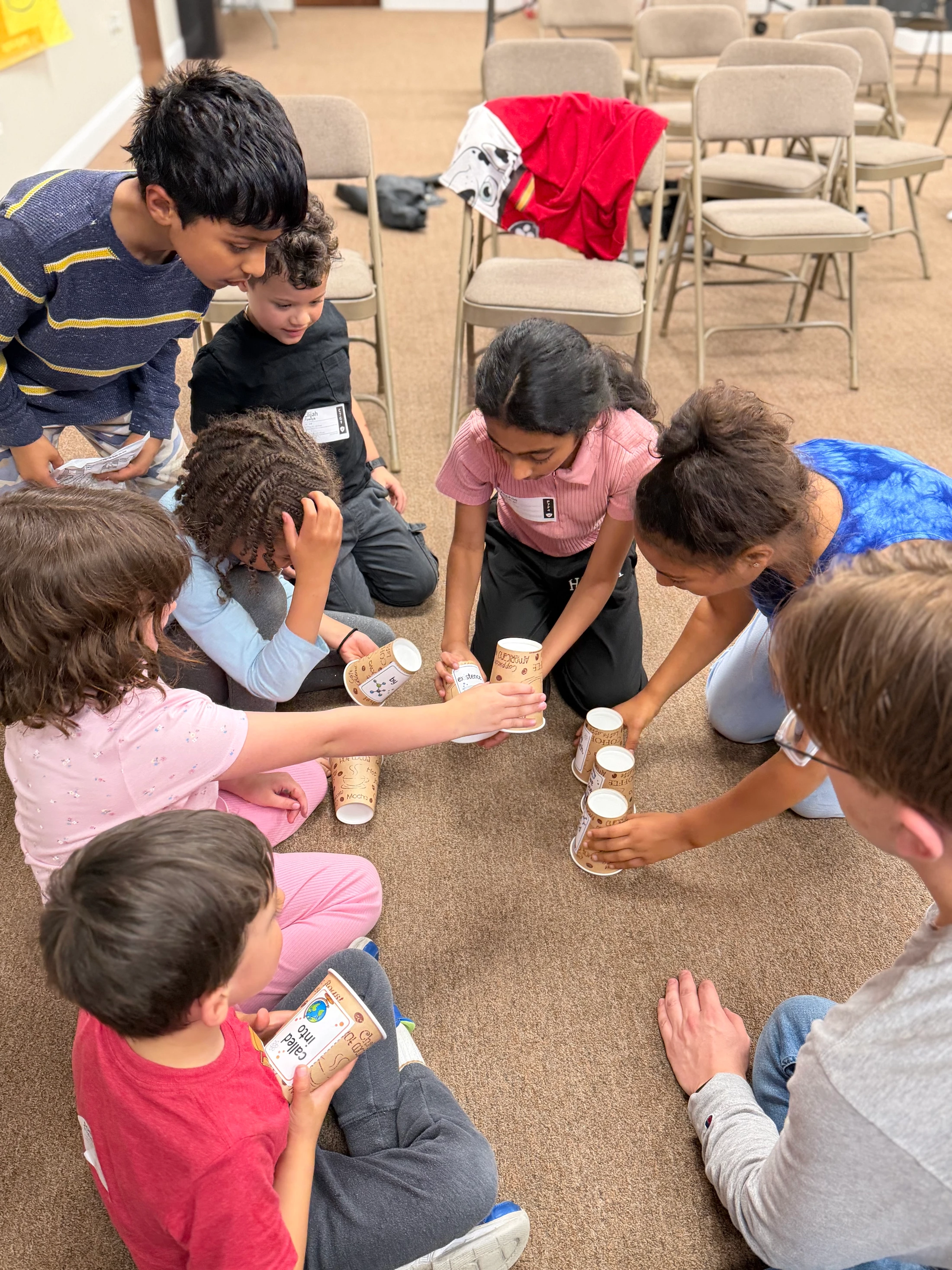 A diverse group of elementary-aged children sit in a circle on a carpeted floor, working together on an activity involving stacking decorated paper cups.