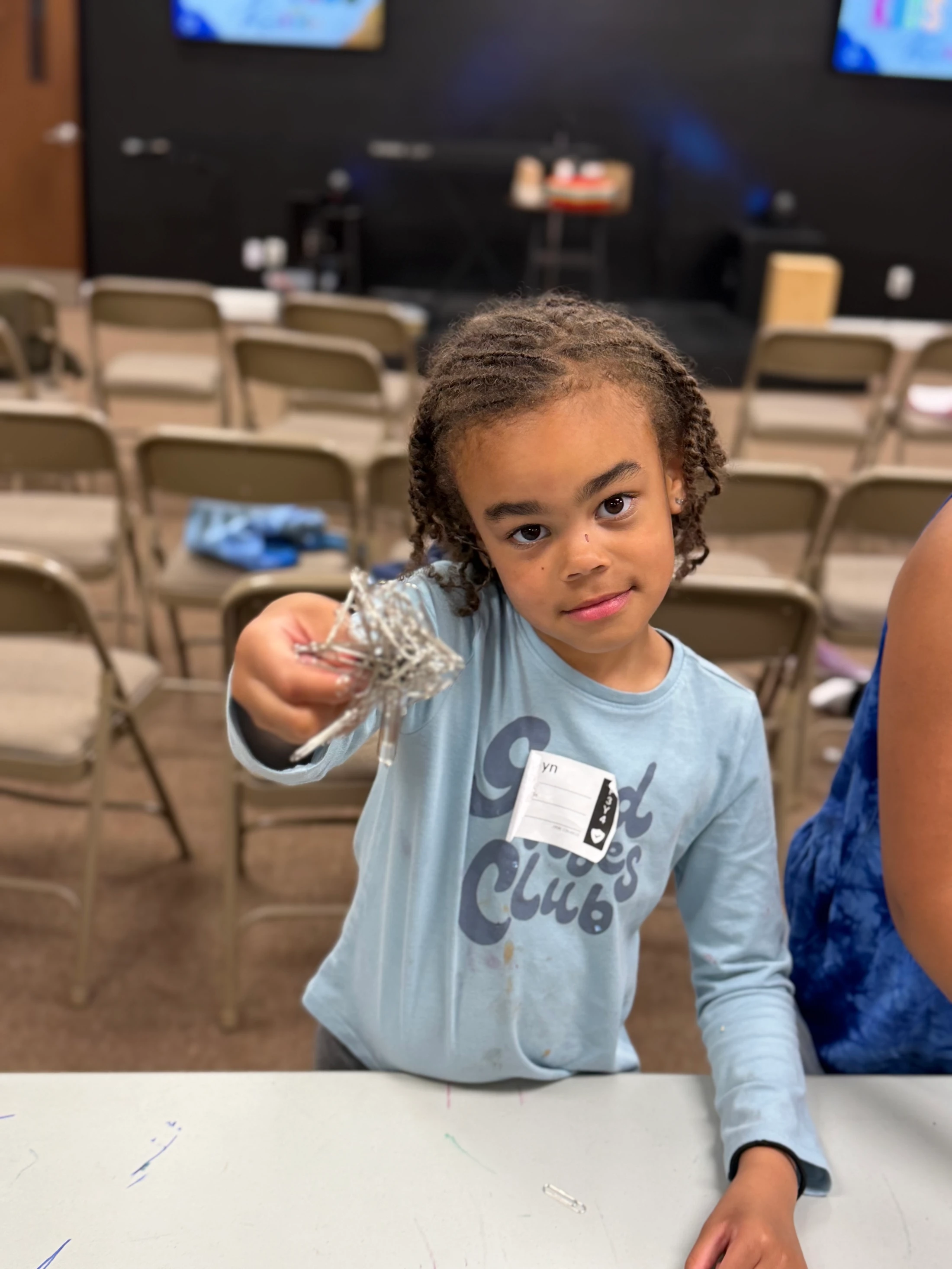 A young girl in a blue shirt with a name tag looks at the camera while holding up a small, tangled silver object she made during an activity.