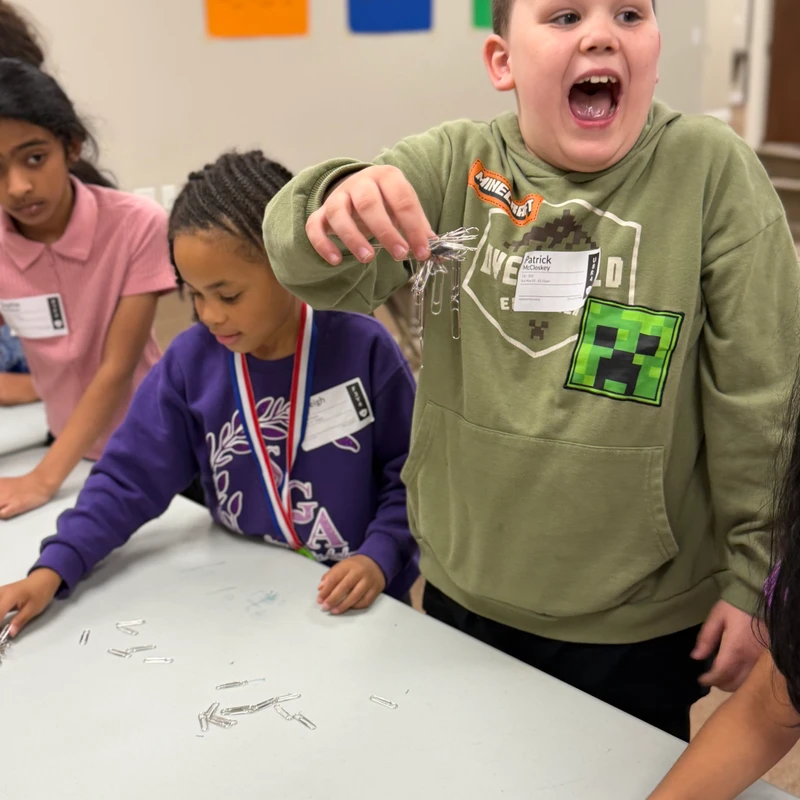 A boy in a green hoodie looks amazed as he holds up a magnet with a cluster of paper clips attached. Two other children watch the experiment at a table.