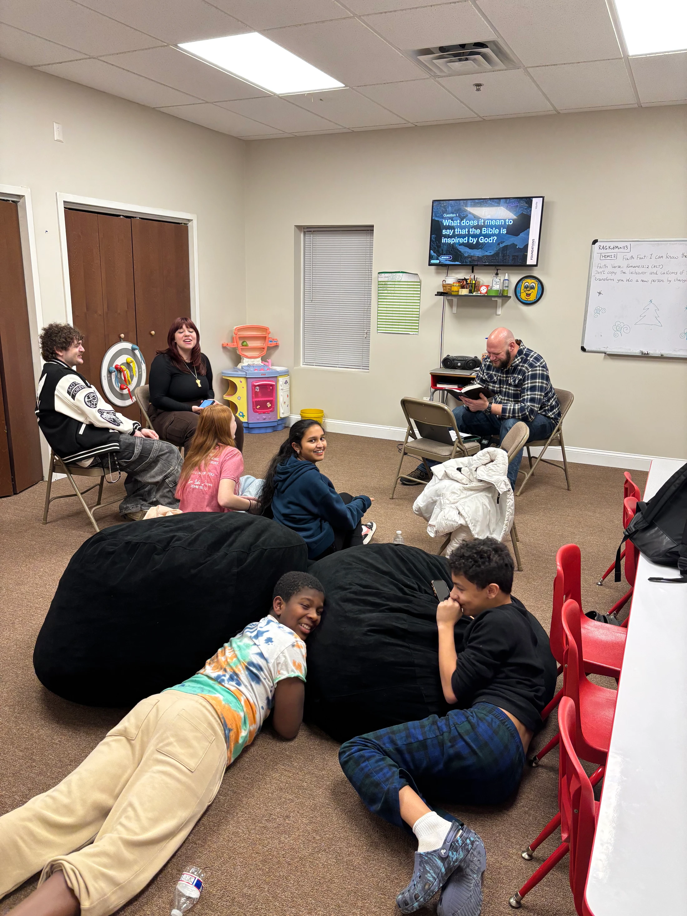 A group of young people and adults are gathered in a classroom for a Bible study, sitting on chairs and beanbags while a man reads from a book.