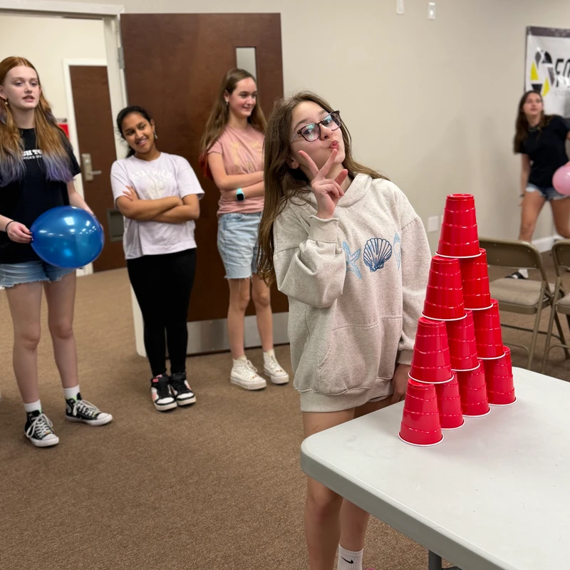 A girl in glasses makes a peace sign next to a pyramid of red cups on a table, while other girls stand in the background at an indoor event.