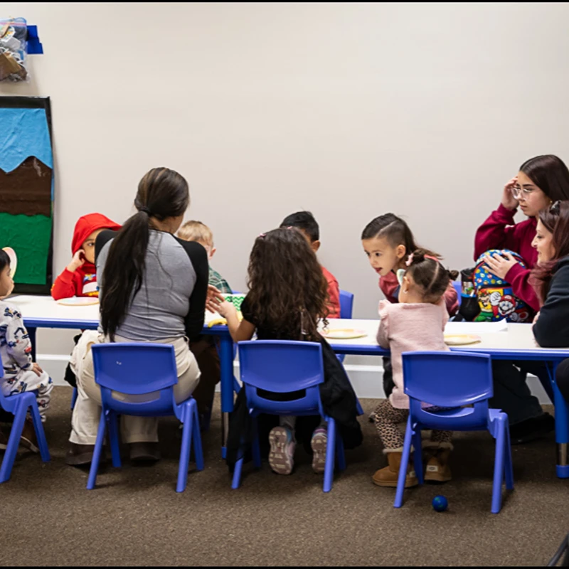 A group of young children and several adults sit together at a long white table with small blue chairs in a classroom.