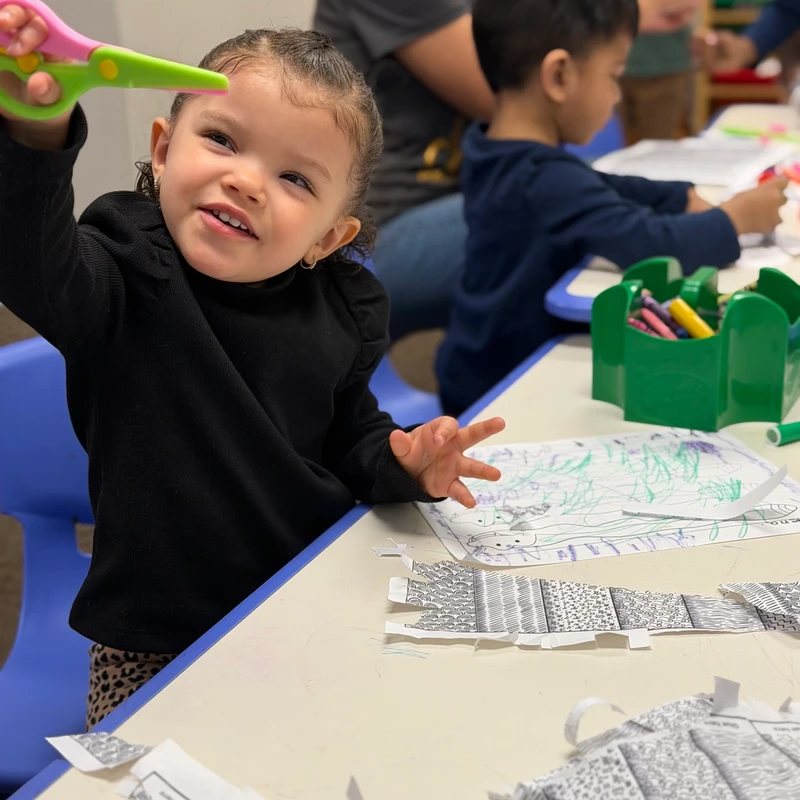 A young girl with her hair in pigtails smiles as she holds up a pair of colorful safety scissors while doing an arts and crafts project at a table.