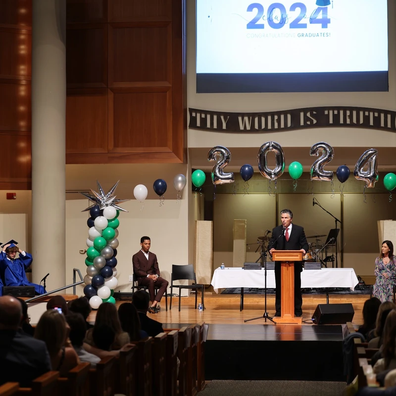 A man in a suit speaks at a podium on a stage decorated with balloons for a 2024 graduation ceremony, with graduates in blue gowns and an audience looking on.