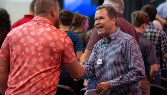 A smiling man in a checkered shirt with a "Dean" nametag shakes hands with another man wearing a red pineapple-print shirt at an indoor event.