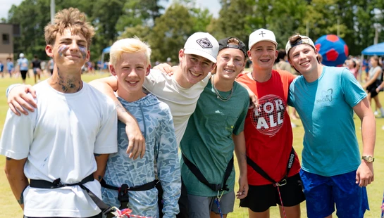 A group of six teenage boys with their arms around each other, smiling for a photo on a sunny day at an outdoor event on a grassy field.