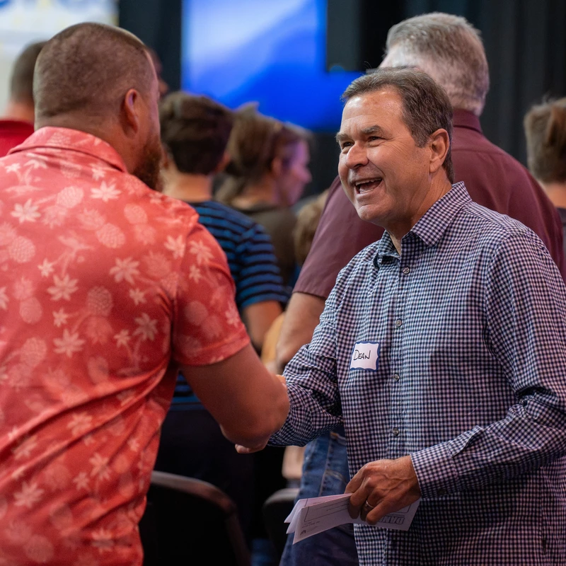 A smiling man in a checkered shirt with a "Dean" name tag shakes hands with another man in a red patterned shirt during an indoor event with other people.