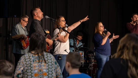 A diverse band performs on a stage with a dark curtain. Men and women sing and play guitars and drums for an audience whose backs are to the camera.