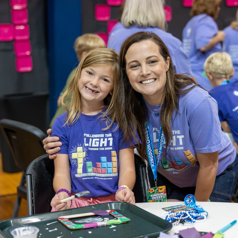 A smiling woman with her arm around a young girl, both wearing matching purple t-shirts, as they sit at a table doing arts and crafts at an event.
