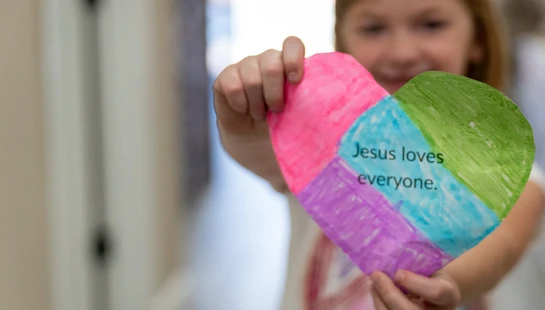 A young girl holds a colorful, hand-colored paper heart towards the camera. The heart reads, 'Jesus loves everyone.'