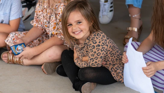 A young girl in a leopard-print shirt sits on the floor with other children, looking at the camera with a happy and engaging smile.