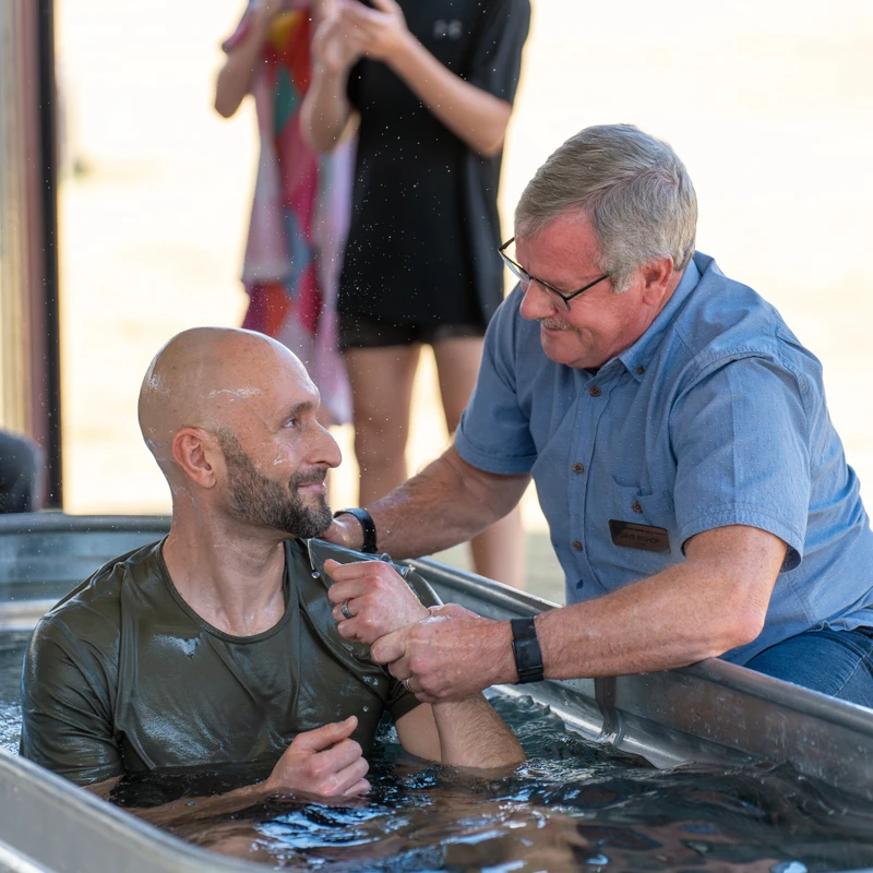 A pastor baptizes a smiling man in a large metal trough of water outdoors. The man being baptized looks up at the pastor during the religious ceremony.