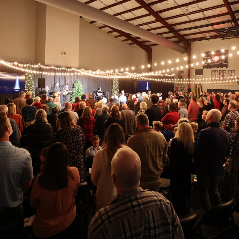 A large crowd stands in a gymnasium decorated for Christmas, watching a band perform on stage under strings of warm lights.