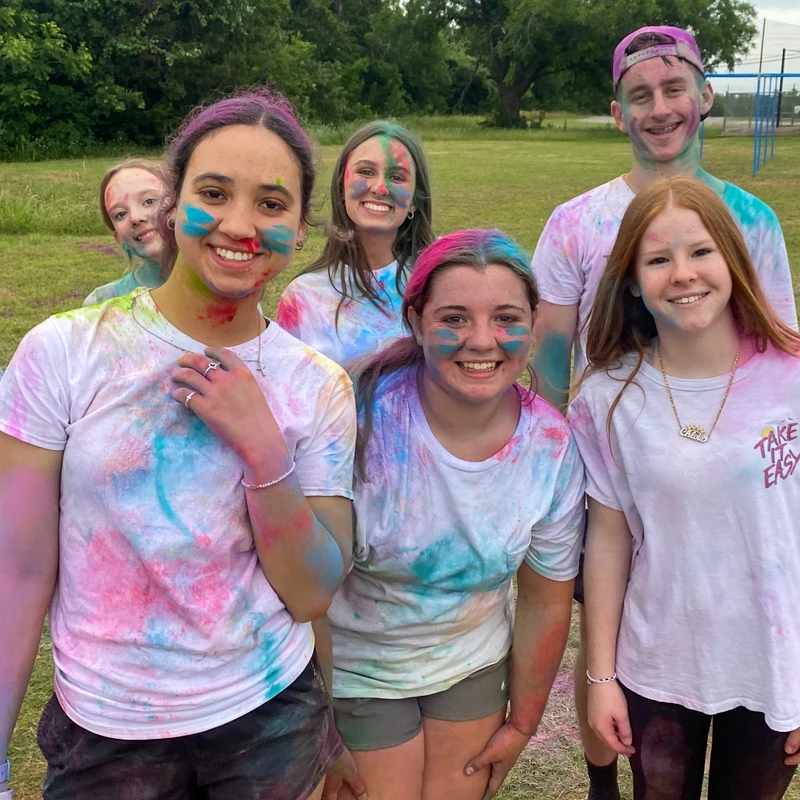 A group of smiling young people at summer camp, their faces and white t-shirts splattered with pink, blue, and purple powder after a color war.