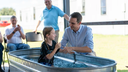A smiling young girl with wet, braided hair sits in a metal tub of water, looking up at a man in a checkered shirt during her outdoor baptism ceremony.