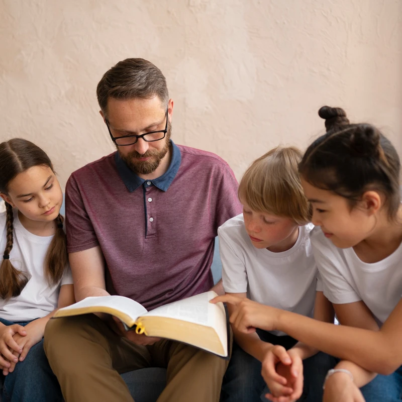 A man with glasses reads from a large open book while sitting on a couch with three children who are looking intently at the page.