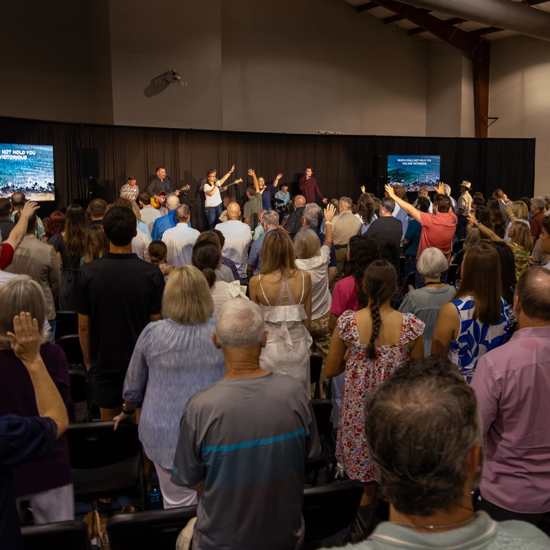 A large congregation stands in a hall, facing a stage where a band leads worship. Many people have their hands raised as they sing along to lyrics on screen.