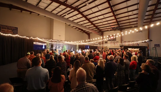 A wide shot from the back of a crowd gathered in a large hall, watching a band perform on a stage decorated with Christmas trees and string lights.