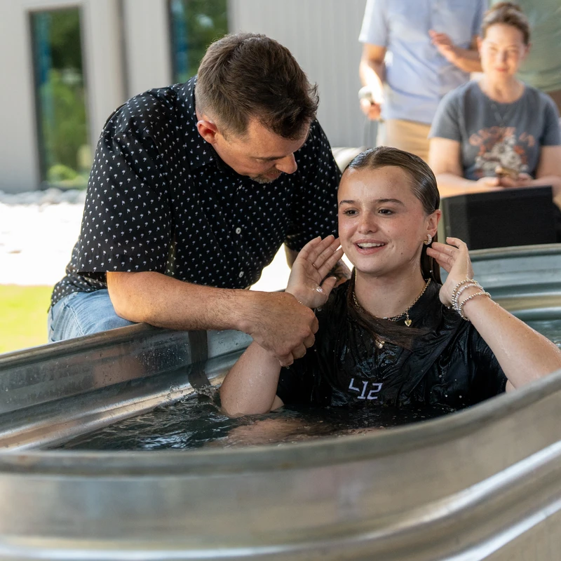 A smiling young woman sits in a metal tub of water during her outdoor baptism ceremony, assisted by a man in a patterned shirt.