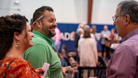 A man in a green shirt with a pen behind his ear smiles while talking to a woman and another man at an indoor social event with people in the background.