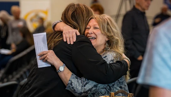 A woman with blonde hair smiles warmly as she hugs another woman at an indoor event. She is holding a paper program in her hand.
