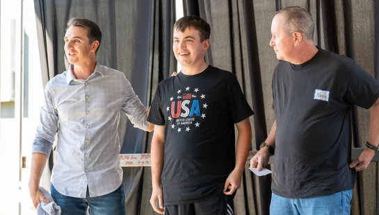 Three men stand together in front of a dark curtain. The man in the middle wears a patriotic USA shirt and smiles as the two other men look on.