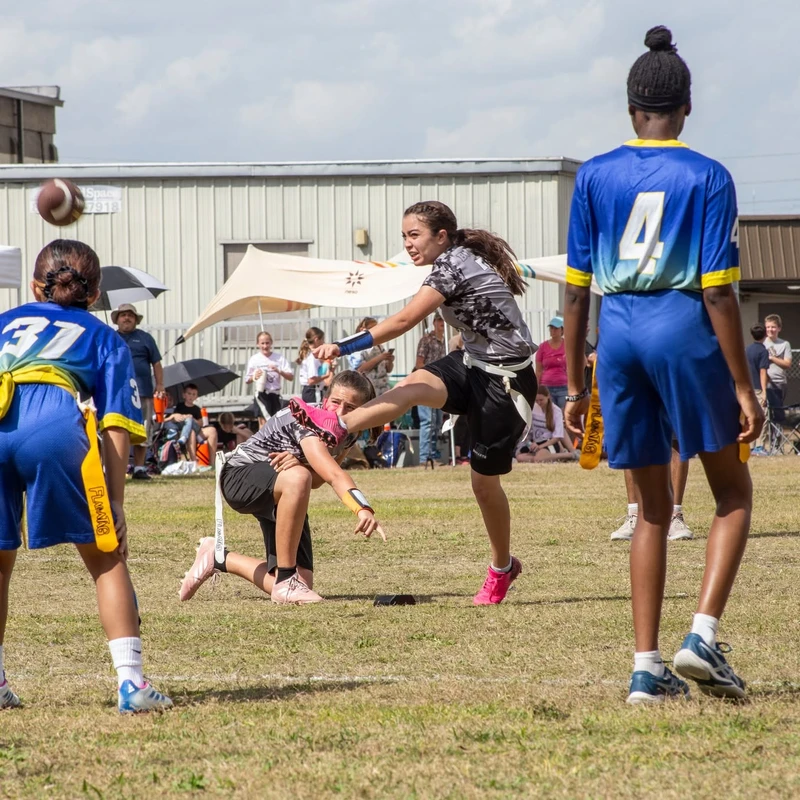 A group of young girls playing flag football on a grassy field. One girl in a grey uniform throws the ball as players from the opposing blue team watch.