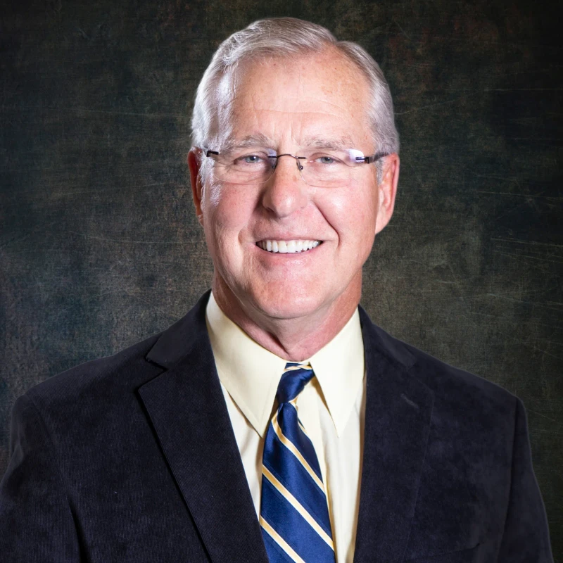 A professional headshot of a smiling older man with gray hair and glasses, wearing a dark suit, yellow shirt, and a blue and gold striped tie.
