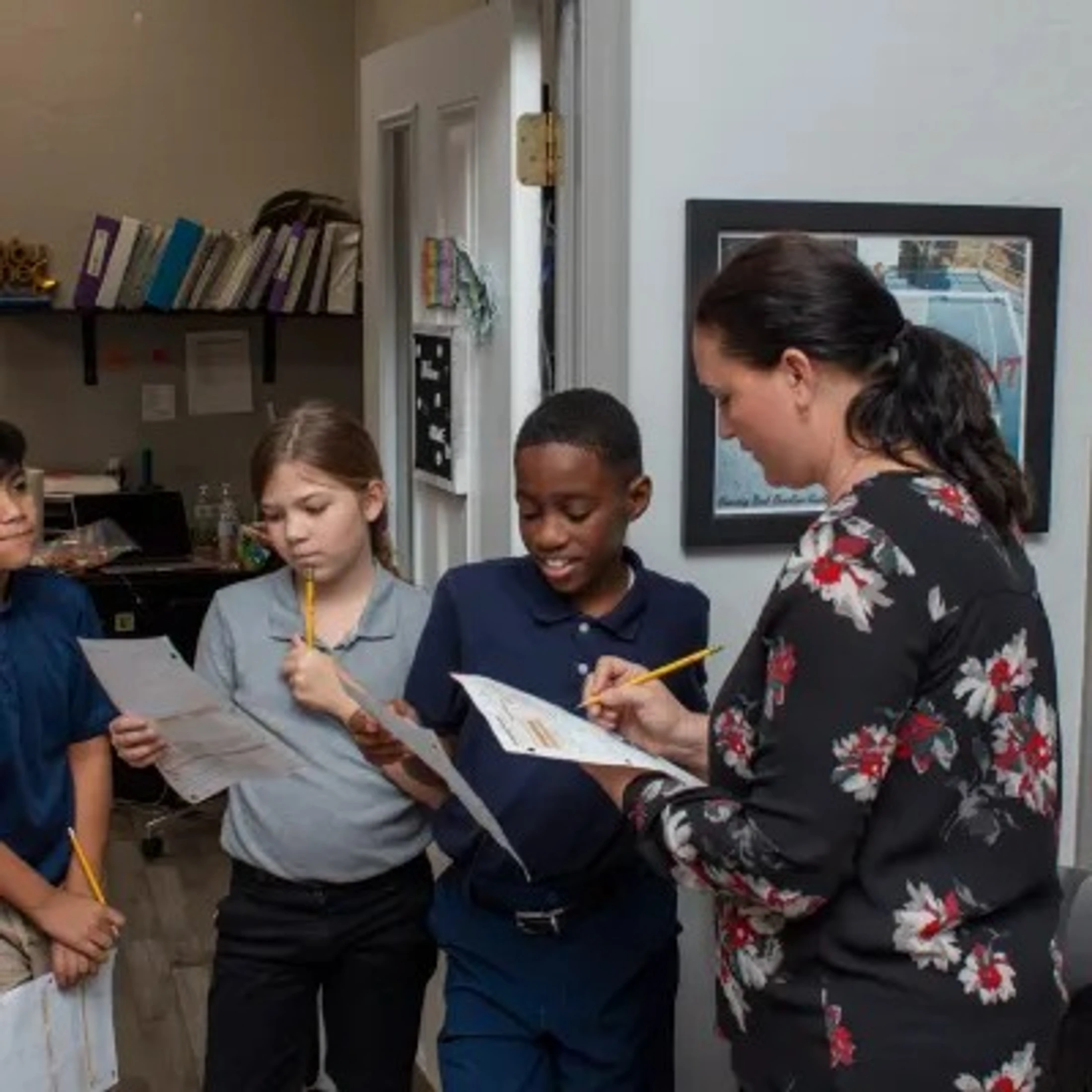 A female teacher wearing a floral shirt stands with three diverse elementary students, pointing to a worksheet one of the boys is holding.