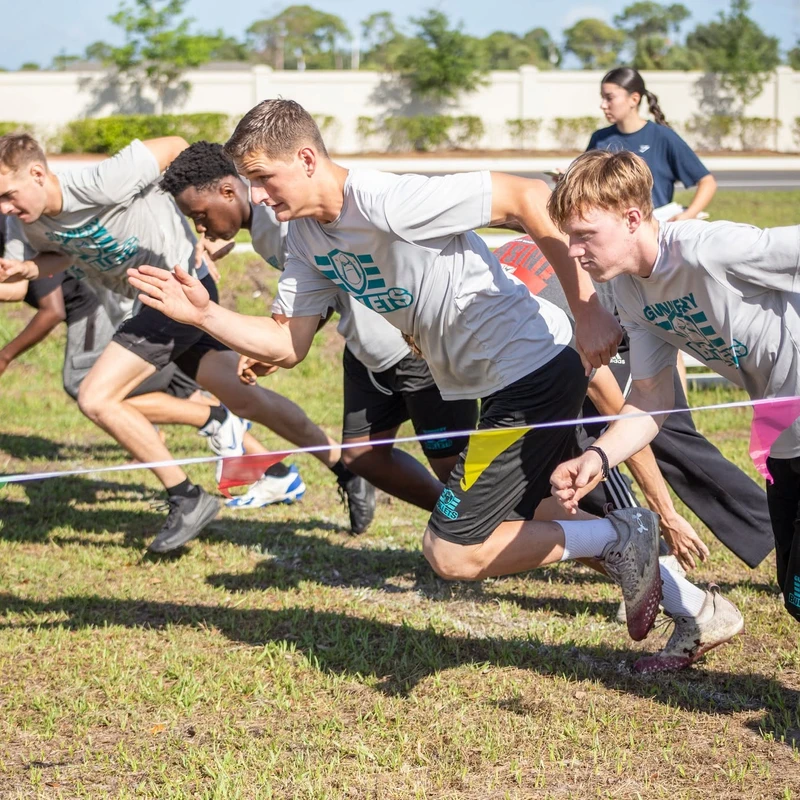 A group of young male runners in athletic gear explode off a starting line on a grassy field during a race on a sunny day.