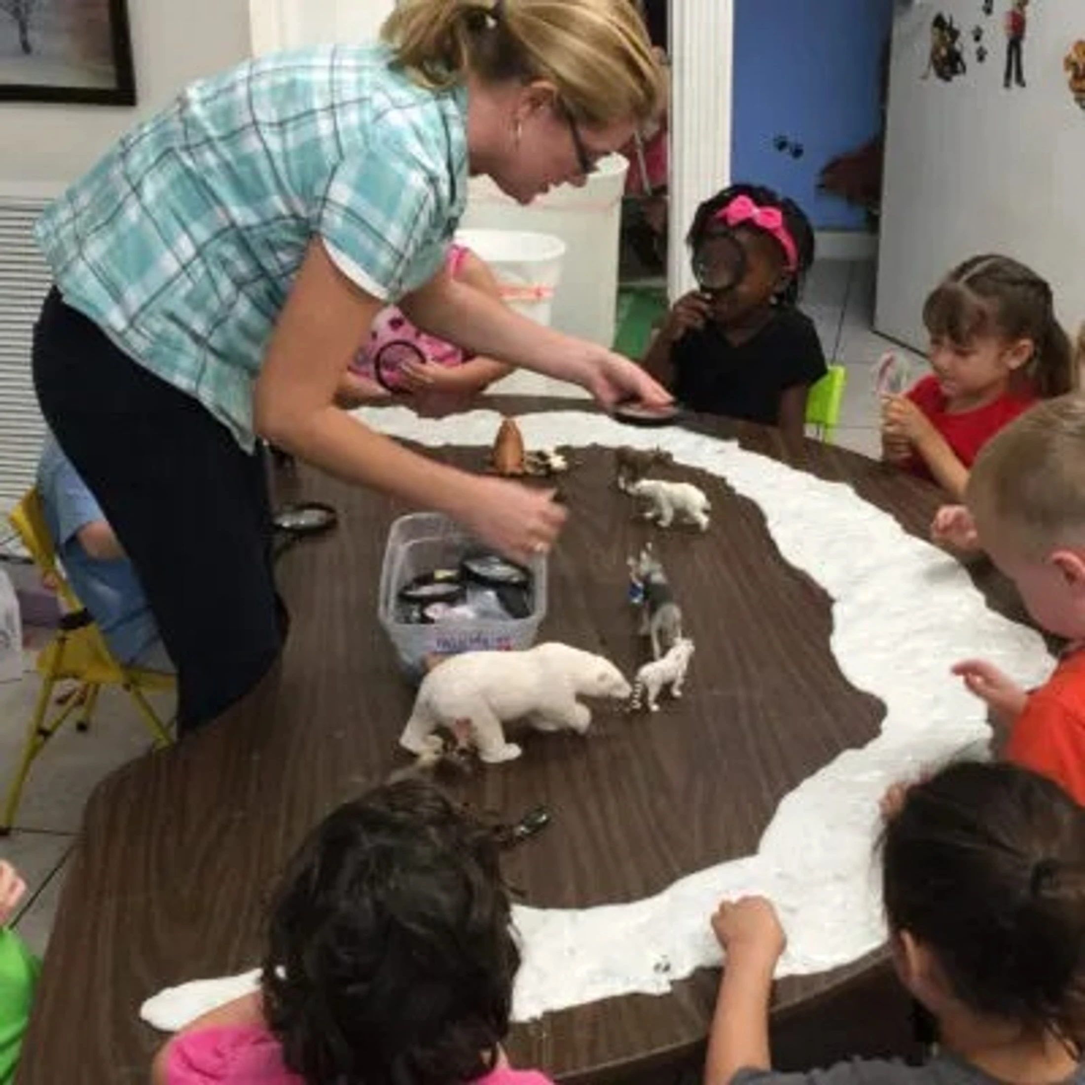 A teacher leads a group of young children in a hands-on activity with toy arctic animals on a table covered with a path of pretend snow.